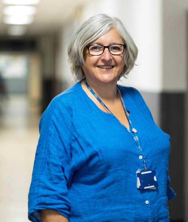 Photo of Professor Caroline Alexander smiling at the camera. She is in a hospital corridor and wearing a blue top.