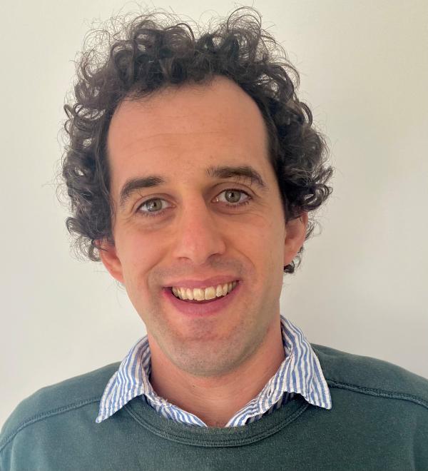 Photo of Dr Peter Hartley smiling at the camera. Peter is wearing a green jumper and a collared shirt.