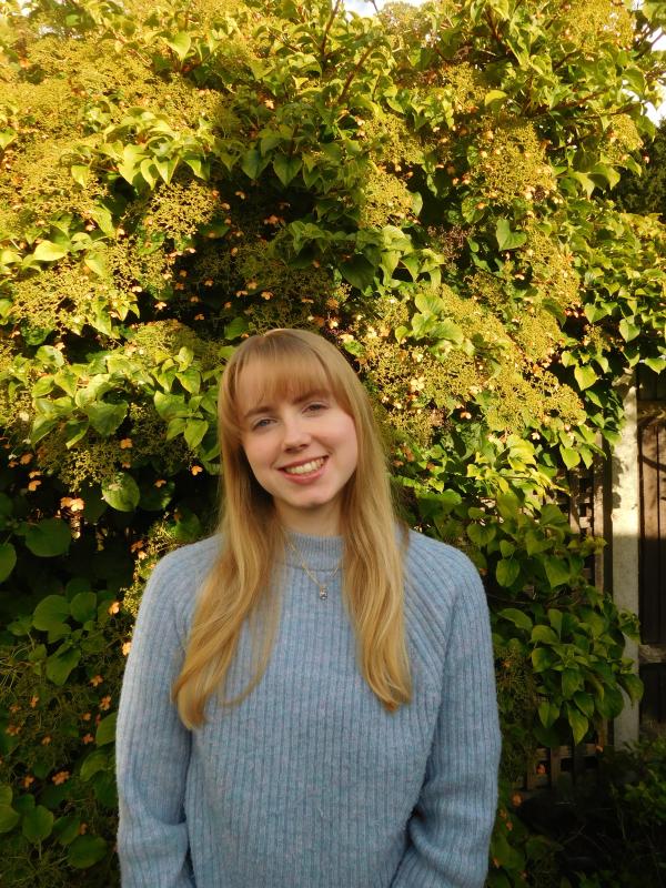A woman with long blonde hair in a light blue jumper in front of a tree