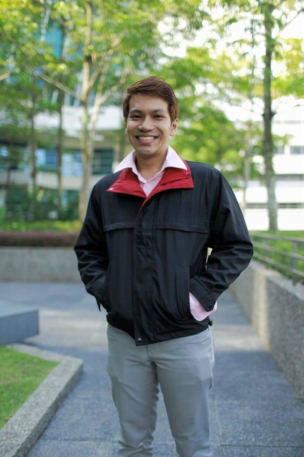 A man standing outside on a path in a black coat with trees behind him