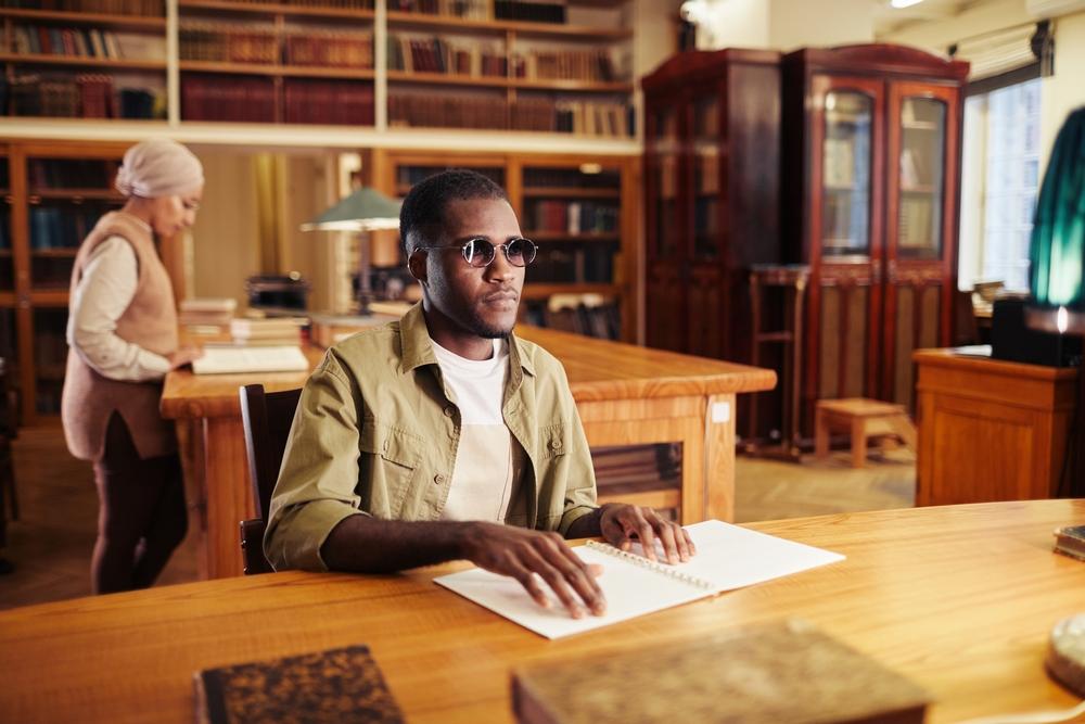 A young black man with vision impairment reading book in braille at table in a university library